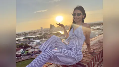Smiling woman in striped jumpsuit and sunglasses sitting on a rooftop at sunset overlooking a city skyline.
