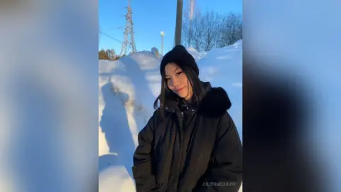 Smiling woman in a black winter puffer jacket and beanie posing in a sunny snowy landscape.