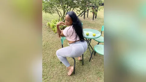 KattyLeya — KattyLeya sits on a green chair outdoors in a pink crop top and tight grey leggings.