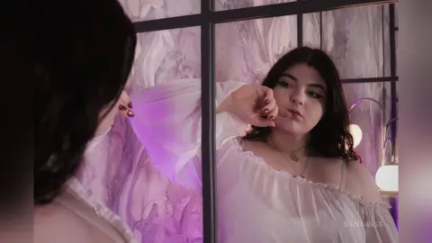 Young woman in a white off-the-shoulder blouse looking into a mirror in a dreamy purple lit room.