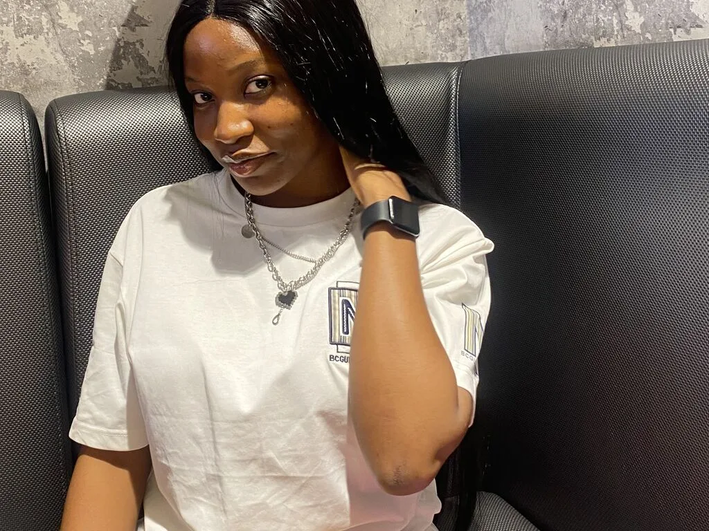 AnnatasiaKath wears a white t-shirt and a silver chain necklace while sitting against a black textured seat.