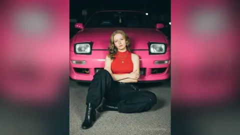 Young woman in a red tank top and black trousers sitting in front of a pink vintage car at night.