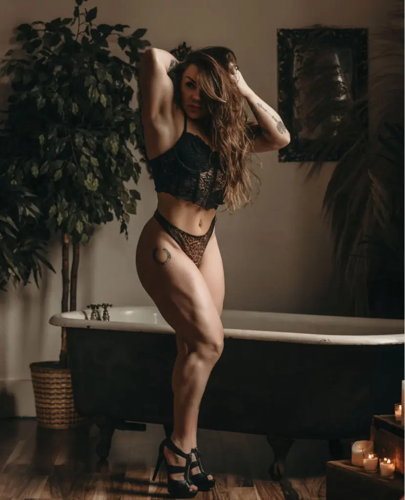 AmaraJezebel poses naked with long brown hair, showing her bare breasts and pussy while standing near a leafy plant and a white bathtub.