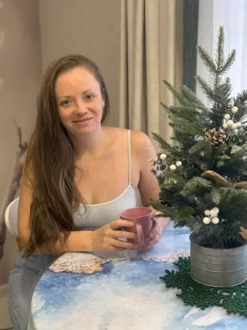 Smiling woman in a white tank top sitting by a small decorated Christmas tree in a cozy indoor setting.