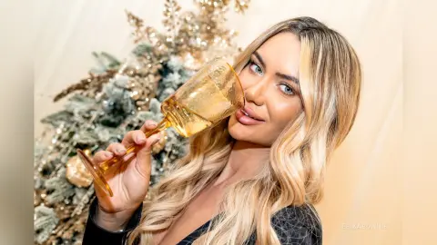 Smiling blonde woman in a black outfit sipping a drink near a festive gold Christmas tree.