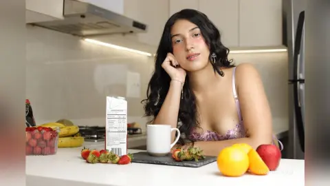 Young woman in a pink lace top sitting at a kitchen counter with fresh strawberries and oranges in a bright mood.