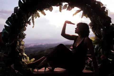 Raven_Morningstar sits in a leafy archway, silhouetted against a sunset view while looking up with her hand near her forehead.