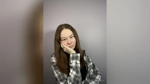 Young woman in a grey plaid shirt leaning her head on her hand against a plain wall with a thoughtful mood.