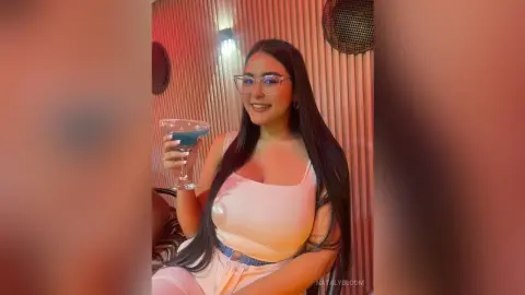 NatalyBloom is a smiling brunette in glasses and a tight white tank top, holding a blue cocktail in a glass.