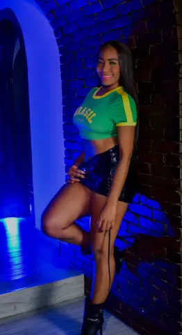 Smiling woman in a green Brasil crop top and black leather shorts posing against a dark brick wall.