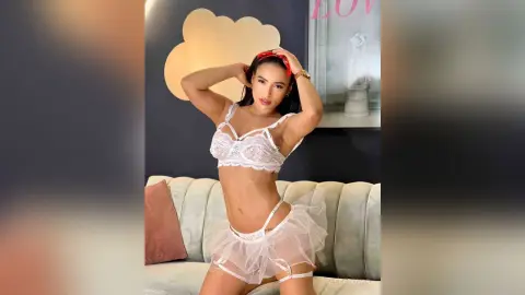 Young woman in white lace lingerie and sheer skirt posing confidently in a modern living room.