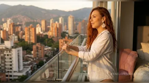 Redheaded woman in a white linen shirt relaxing on a high-rise balcony overlooking a city skyline at sunset.
