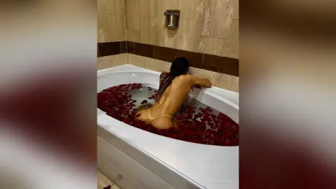 Nude woman relaxing in a white bathtub filled with red rose petals in a luxury bathroom setting.