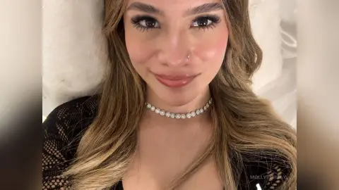 Smiling young woman with long wavy hair wearing a black lace top and sparkling diamond choker in a cozy setting.