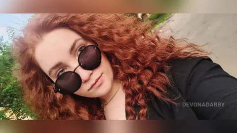 Redheaded woman in black outfit and round sunglasses smiling outdoors in natural sunlight.