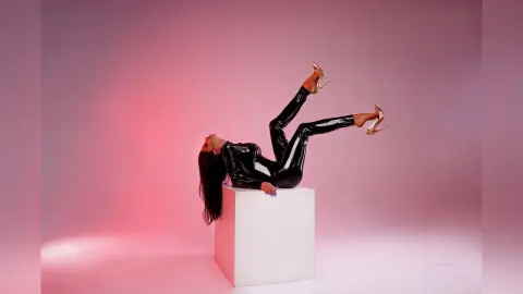 Fashionable woman in a black shiny bodysuit and gold heels reclining on a white pedestal against a pink backdrop.