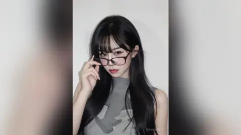 Young woman with long black hair and glasses wearing a grey sleeveless top in a minimalist studio setting.