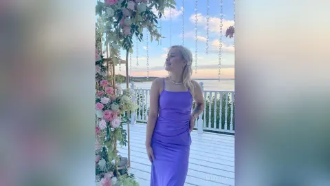 EmilyGiffin smiles while wearing a tight purple tank dress, standing on a white deck decorated with pink and white flowers.