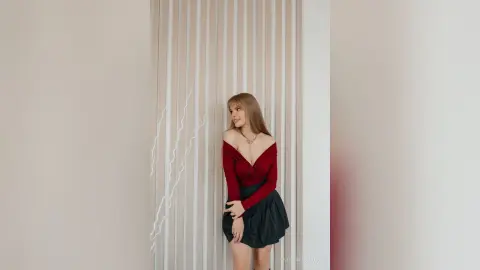 Young woman in a red off-the-shoulder top and black skirt posing against a minimalist striped wall.