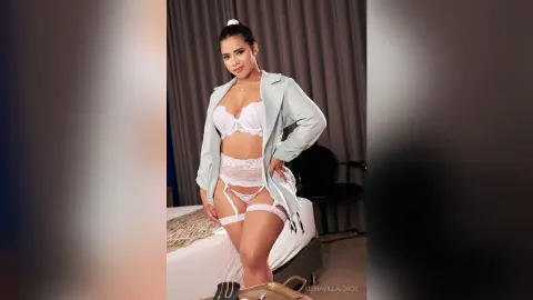 Young woman in white lace lingerie and a light mint blazer posing confidently in a modern bedroom setting.