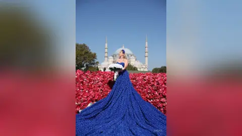 Elegant woman in a long blue gown holding a white bouquet in front of a grand white mosque under a clear sky.