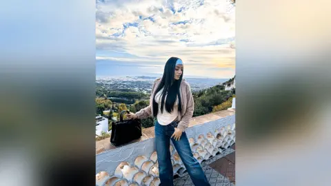 Aleja stands on a balcony overlooking a city view, wearing a light tan top, a brown jacket, and blue jeans.