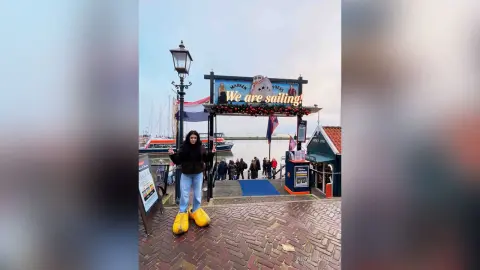 AnnaBanks stands outside wearing a black jacket and light blue jeans with bright yellow boots on a brick walkway near a harbor sign.