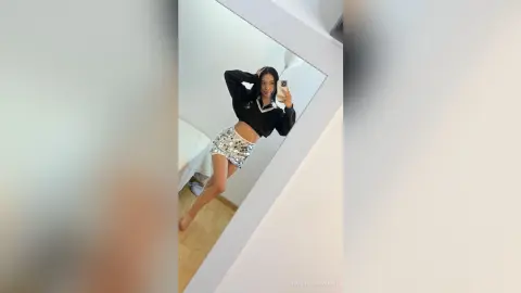 Stylish young woman in a black crop top and silver sequin mini skirt taking a mirror selfie in a bright room.