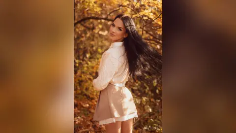 Beautiful woman in a white blouse and beige skirt posing in a golden autumn forest setting.