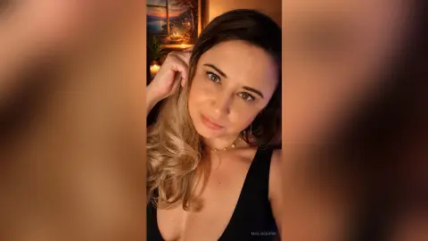 MaliaQuinn poses with her hand near her face, showing her blonde hair and black tank top in a warm, indoor setting.