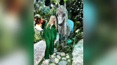 Blonde woman in emerald green silk pajamas standing next to a magical unicorn statue in a lush garden setting.