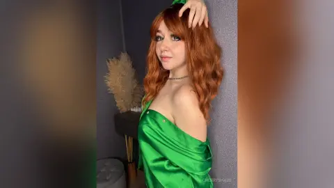 Young woman with wavy red hair wearing a green satin dress in a modern indoor setting.