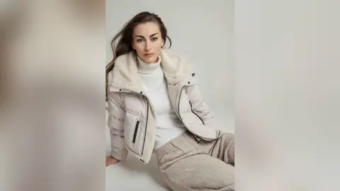 Young woman in a cozy white turtleneck and cream puffer jacket posing in a bright, minimalist studio setting.