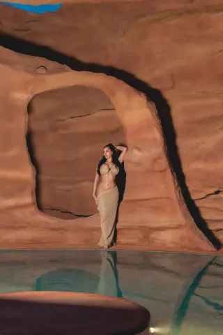 Elegant woman in a beige bikini and long skirt posing in a dreamy desert cave by a turquoise pool.