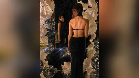 KimRece poses with her back turned, wearing a black backless dress, standing in front of a mirror surrounded by white roses and disco balls.