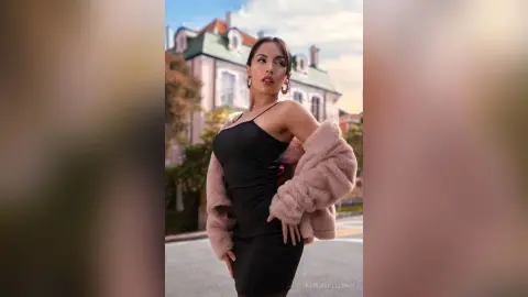 Elegant woman in a black satin dress and pink faux fur coat posing outdoors near a classic building.