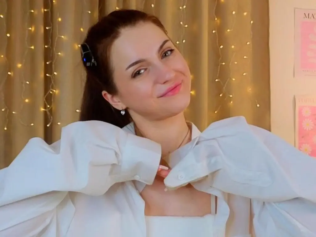 Smiling young woman in a white button-down shirt posing in a cozy room with warm fairy lights.