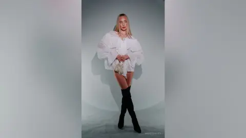 MarryDavies stands against a grey background wearing a white puffy-sleeved blouse and black thigh-high boots.