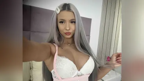 Kalira poses in a white lace bra and pink trim, showing off her long silver hair and cleavage while sitting against a grey headboard.