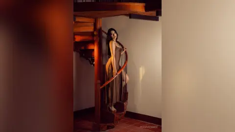 Elegant woman in a brown sleeveless dress posing on a wooden spiral staircase in warm, moody lighting.