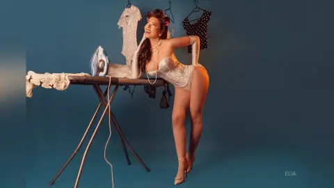 Glamorous woman in white lace lingerie and fishnets leaning over an ironing board in a blue studio setting.