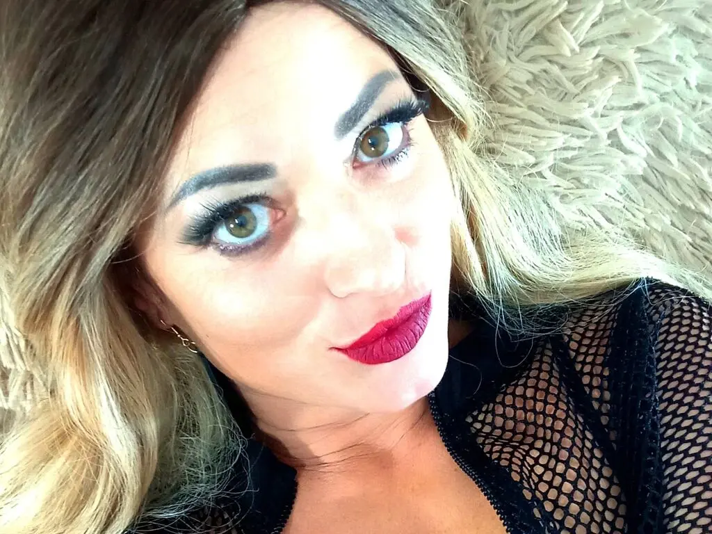 Smiling blonde woman in a black fishnet top lying on a soft white shag rug for a glamorous close-up portrait.