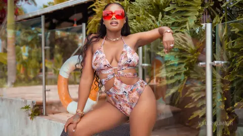 Curvy woman in a patterned cutout swimsuit and red sunglasses posing outdoors in a tropical garden setting.