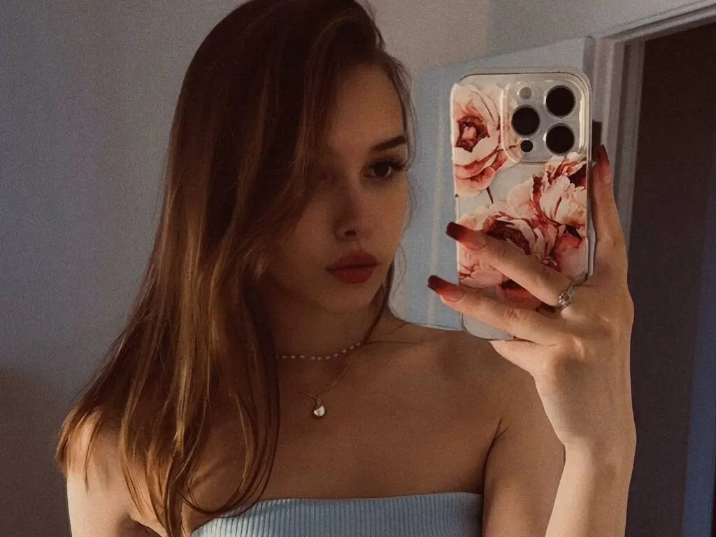 TarenLeonetti poses for a mirror selfie wearing a light blue tube top and a pearl necklace, showing her long brown hair and red nails.