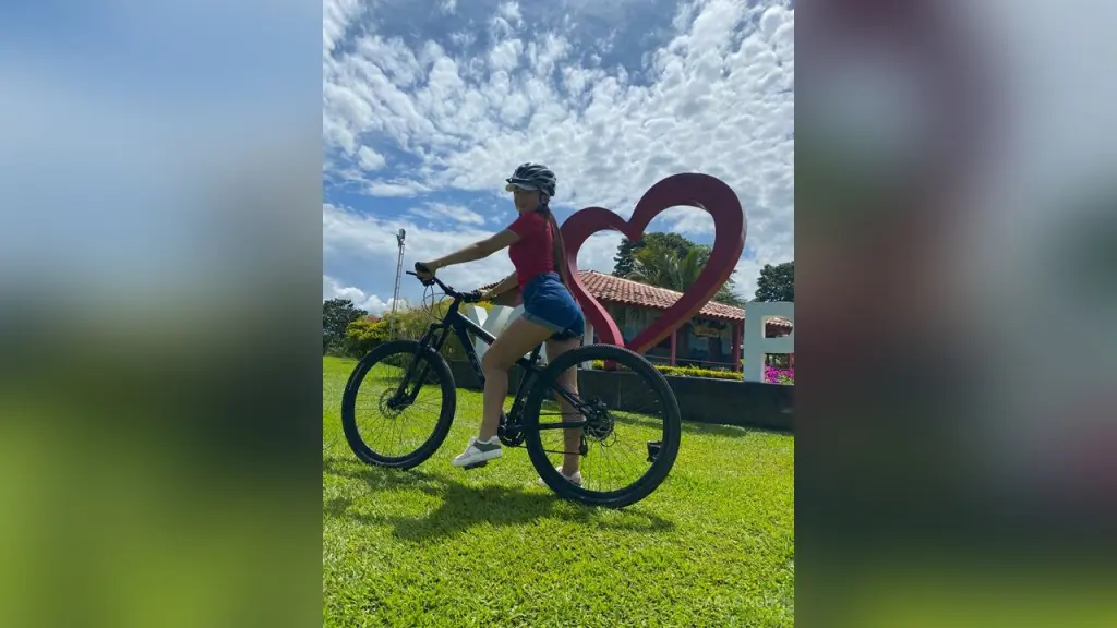 SaraSingh — Young woman in red shirt and denim shorts riding a bicycle past a large red heart sculpture on a sunny day.