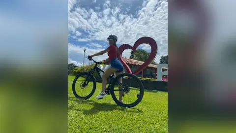 Young woman in red shirt and denim shorts riding a bicycle past a large red heart sculpture on a sunny day.