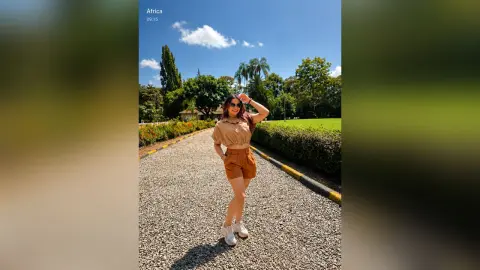 SofiaGreco stands on a gravel path wearing a tan crop top, matching shorts, and white sneakers under a bright blue sky.