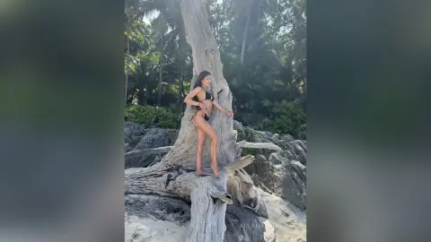 MilanaVolkova poses in a black bikini while standing on a large piece of driftwood on a tropical beach surrounded by palm trees.
