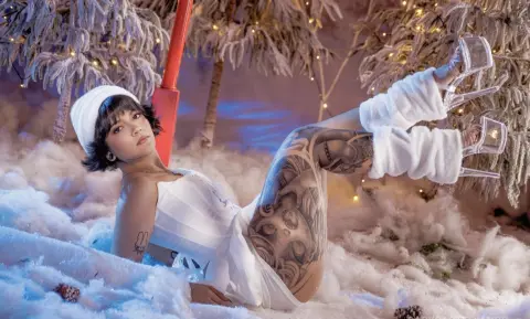 Tattooed woman in white winter outfit lounging in a snowy wonderland setting with a dreamy mood.