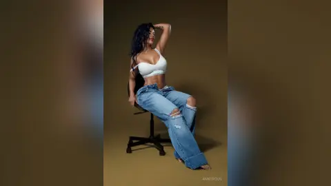 AnnyRoug poses sitting on a black chair wearing a white bra and ripped blue jeans, showing her midriff and long dark hair.